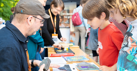 Zwei Kinder stehen vor einem Tisch und schauen sich Ausstellungsstücke an, während ein Mann ihnen etwas erklärt