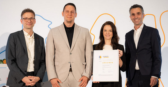 Vier Personen posieren für ein Foto. Eine hält eine große Urkunde.