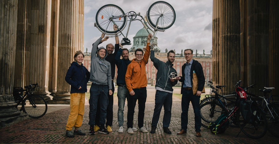 Gruppe von Menschen steht zwischen Säulen, einer übergibt ihnen einen Pokal