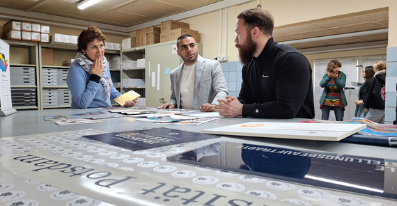 Drei Personen sind um einen Tisch versammelt auf dem Druckerzeugnisse ausgelegt sind.