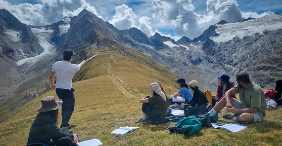 People sitting at a mountain ridge, one person is standing in front of them, explaining while gesturing.