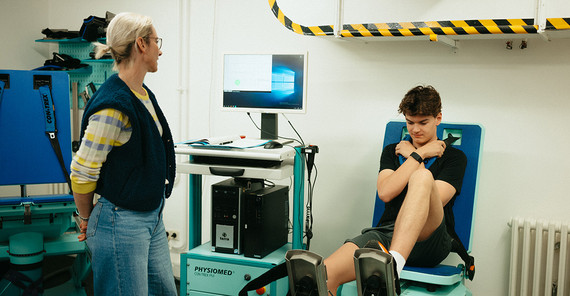 Dr. Josefine Stoll mit Flynt Schuring Eine blonde Frau schaut auf einen Monitor. Auf einem Sitz neben ihr sitzt ein junger Mann mit dunklen Haaren, ein Bein auf dem Boden, eins auf einem Pedal, die Arme vor der Brust gekreuzt.