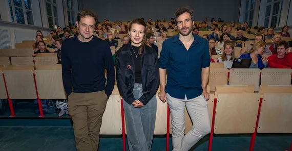 Drei Personen stehen mit dem Rücken zum Auditorium eines gut gefüllten Hörsaal und posieren für das Foto.