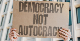 Ein Pappschild wird hochgehalten. Daruaf zu lesen: democracy not autocracy