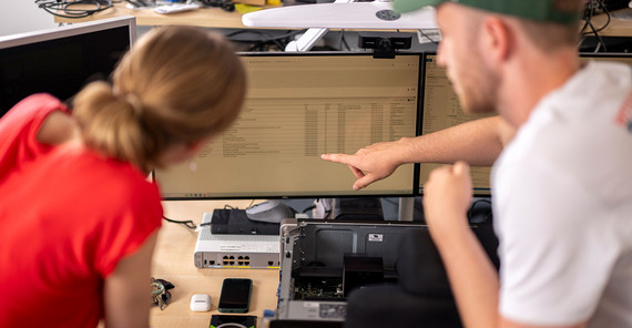Two people can be seen from behind. They are sitting in front of a computer screen. One is explaining something to the other by pointing at it.