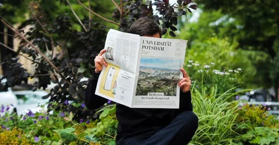Auf dem Foto sieht man einen schwarz gekleideten Mann, der die neueste Ausgabe der Beilage im Park liest.