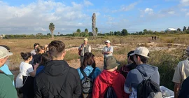 Gruppe von Menschen stehen vor einer Ausgrabungsstätte, während ihnen ein Mann etwas erklärt