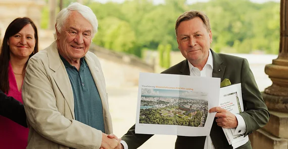 Zwei Männer schütteln einander die Hand, einer hält einen Ausdruck eines Bauentwurfs in die Kamera