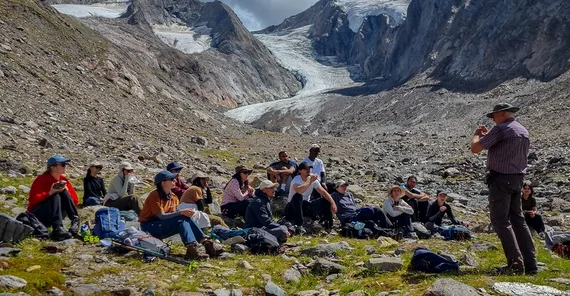 Menschen sitzen vor einer Berglandschaft, während einer vor ihnen steht und etwas erzählt