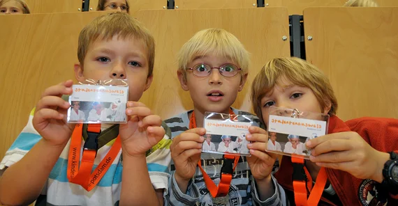 Drei blonde Kinder in einem Hörsaal zeigen ihre Studierendenausweise