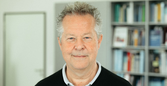 Prof. Dr. Frank Mayer Ein Mann mit grauen Haaren in einem schwarzen Pullover. Im Hintergrund sieht man unscharf ein Bücherregal.