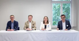 Vier Personen sitzen an einem Tisch und schauen in die Kamera