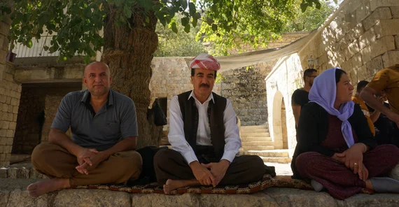 Das Bild zeigt im Vordergrund drei Personen die auf einer Mauer sitzen.