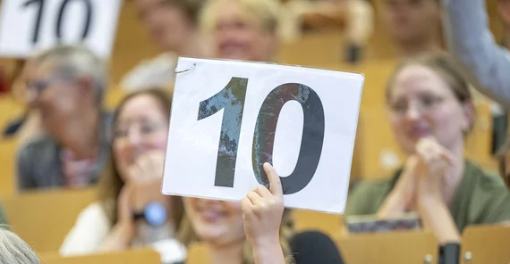 Hand, die ein Schild mit einer 10 darauf hochhält, im Hintergrund Menschen in einem Hörsaal.