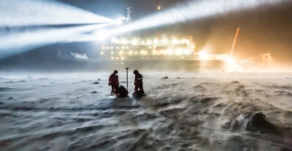 Dramatische Szenerie einer Polarregion bei Nacht: Das Forschungsschiff "Polarstern" im Hintergrund erleuchtet Schnee und Eislandschaft im Vordergrund, wo zwei Menschen arbeiten.