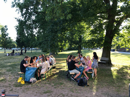 Zwei Bierbänke auf einer Wiese mit Bäumen, an denen Studierende sitzen, die essen und trinken.