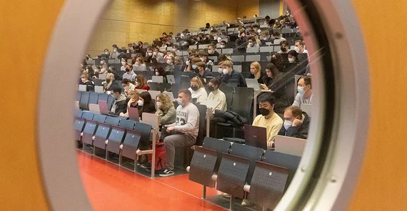 Blick durch das Bullauge in Hörsaal 5 in Griebnitzsee. Studierende sitzen mit Maske in den Reihen.