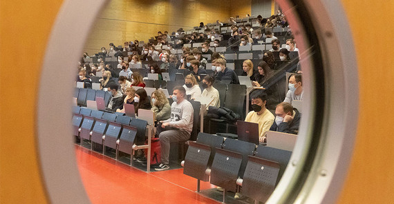 Blick durch das Bullauge in Hörsaal 5 in Griebnitzsee. Studierende sitzen mit Maske in den Reihen.