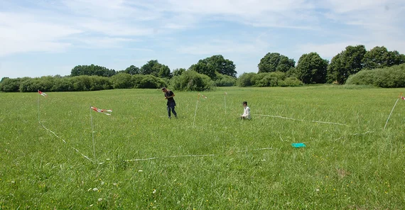 Bild von einer Wiese, auf der ein Stück mit Flatterband abgesperrt ist, darauf zwei junge Menschen