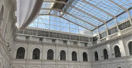 Ein Blick nach oben in das gläserne Dach des Archäologischen Museums durch welches blauer Himmel zu erkennen ist. Die Metall-Glaskonstruktion wird getragen von weißen Wänden, die rhythmisch auf mehreren Etagen durch Torbögen unterbrochen sind.