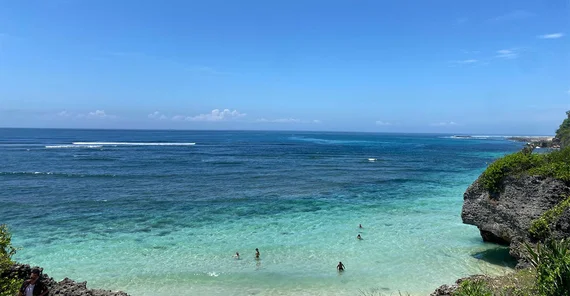Strand auf der Halbinsel Bukit. Das Meerwasser ist am Strand hell-türkis und wird dann immer dunkler und blauer. Das Meer reicht bis zum Horizont. Am Strand befinden sich einige badende Menschen.