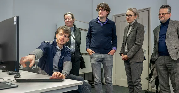 Eine Gruppe von Menschen steht in einem Raum vor einem Computer, während einer etwas erläutert.