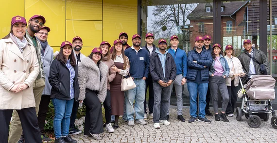 Die Teams des aktuellen Impact Labs an der Universität Potsdam bestehend aus gut einem Dutzend Menschen, die vor einem gelben gebäude posieren.