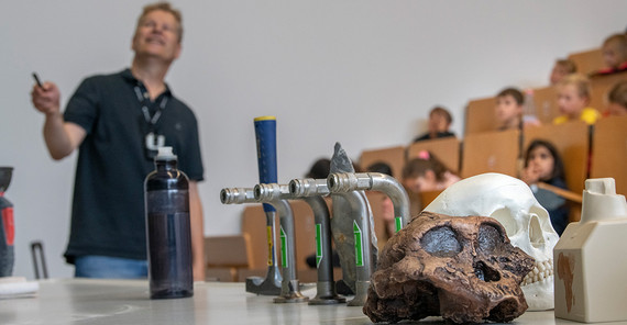 Ein Professor zeigt etwas, vor ihm auf dem Tisch liegen zwei Schädel. Im Hintergrund sieht man Kinder in einem Hörsaal.