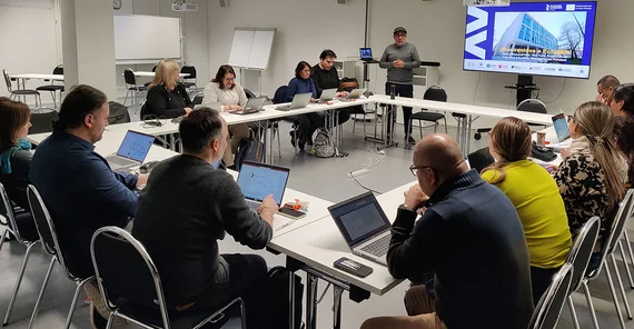 Menschen in einem Konferenzraum an Tischen sitzend, die im Rechteck aufgebaut sind. Eine Person referiert vor einer digitalen Anzeige.