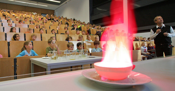 Eine rote, helle Flamme kommt aus einem Porzellangefäß. Im Hintergrund erklärt Prof. Holdt etwas dazu, Kinder im Hörsaal schauen zu.