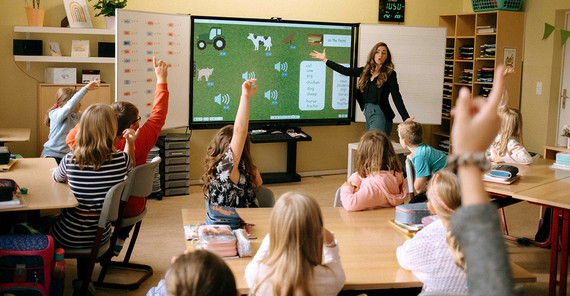 Eine Lehrerin steht vor einer Klasse und erklärt. Viele Kinder melden sich.