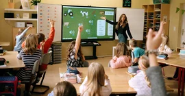 Eine Lehrerin steht vor einer Klasse und erklärt. Viele Kinder melden sich.